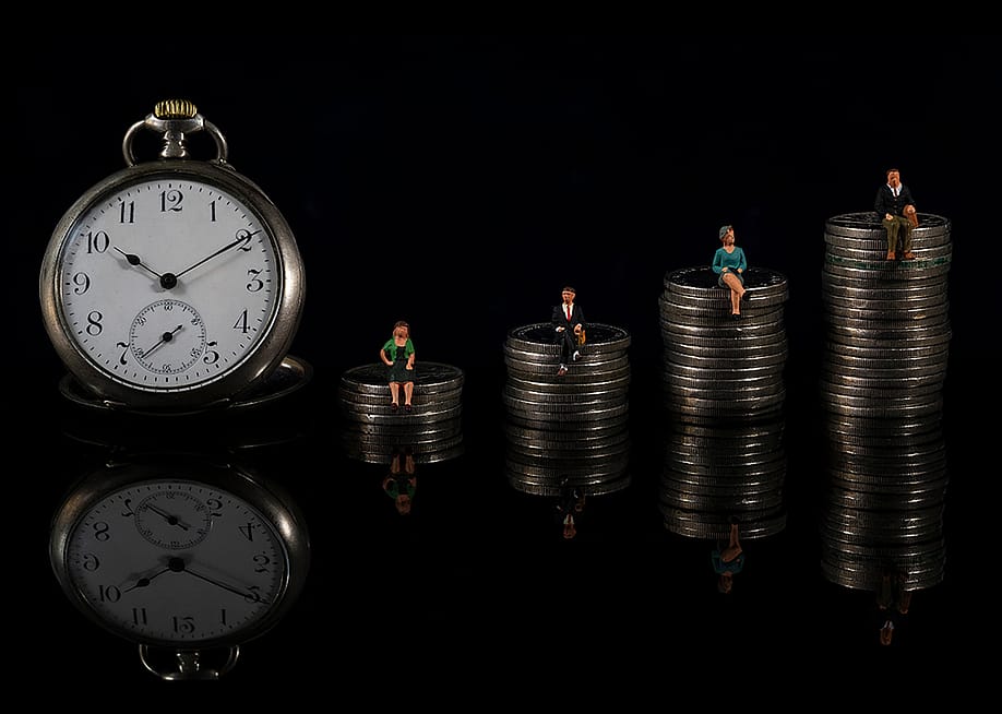 The concept of counting down to retirement illustrated by an increasing stack of coins with miniature people sitting on each one, with a clock placed on the side. — pabloavanzini // Shutterstock