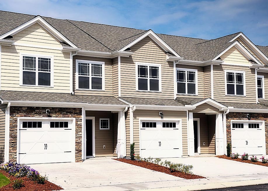 A row of modern townhomes with beige siding, white trim, and stone accents, each featuring a garage, driveway, and small landscaped front yards under a bright blue sky. — Rare pixels // Shutterstock