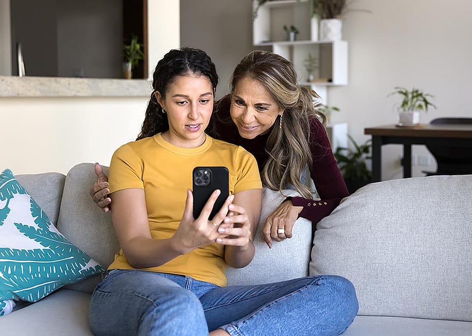 Adolescent daughter on sofa shows interested mum social media on phone screen. — fizkes // Shutterstock