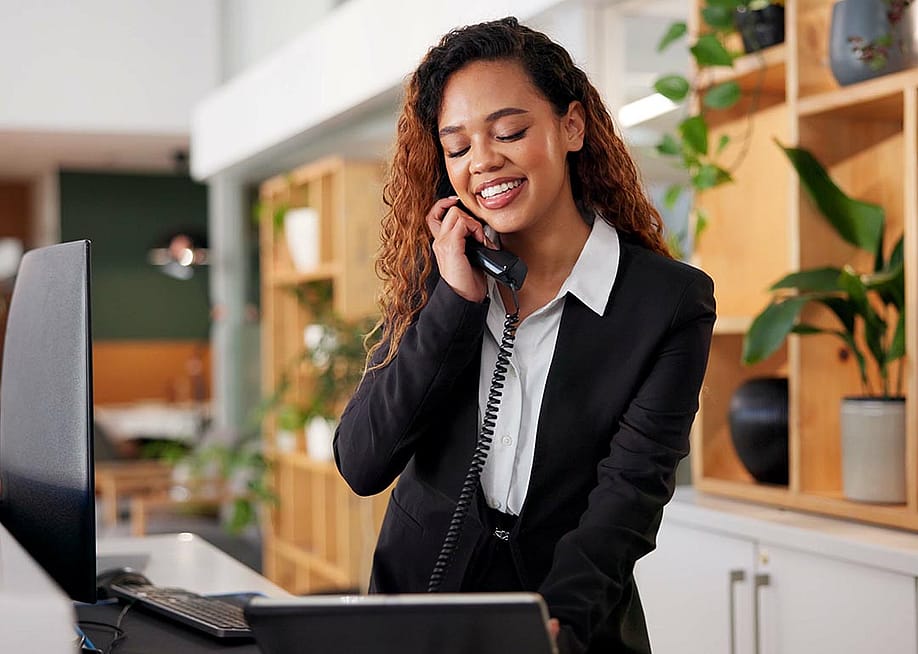 Customer service concierge answering phone standing behind reception desk looking at computer screen with modern decor and houseplants behind her. — PeopleImages // Shutterstock