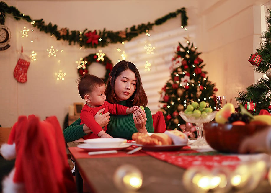 A young mother sitting alone with holding her baby at the dinner table during Christmas Eve. — Panya_photo // Shutterstock