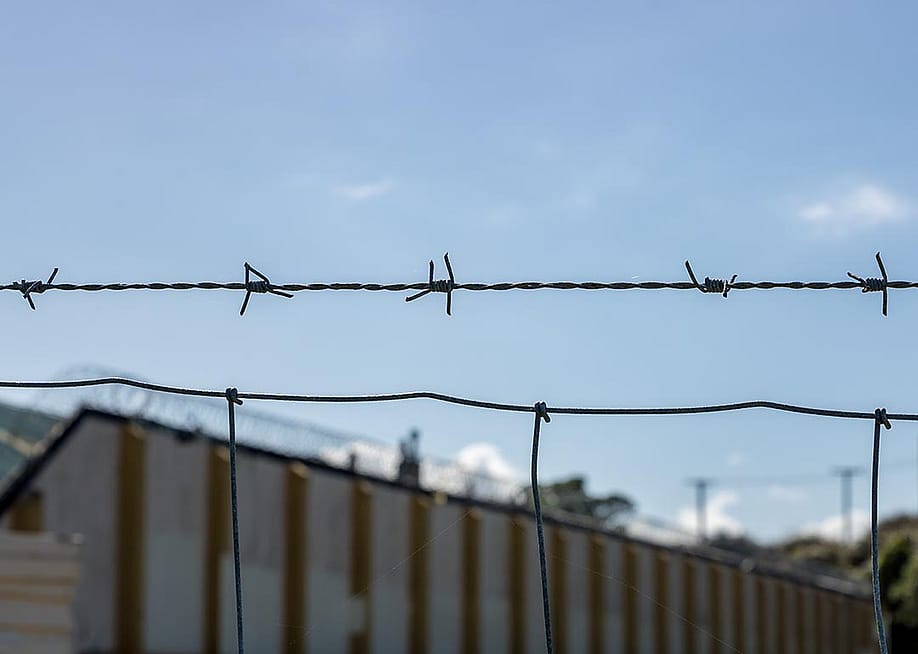 Close up of a single strand of barbed wire above a fence with a correctional facility in the background. — JoshuaDaniel // Shutterstock