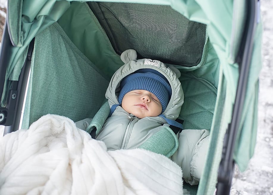 A baby in winter clothes sleeping in a green stroller outdoors. — Jayakri // Shutterstock