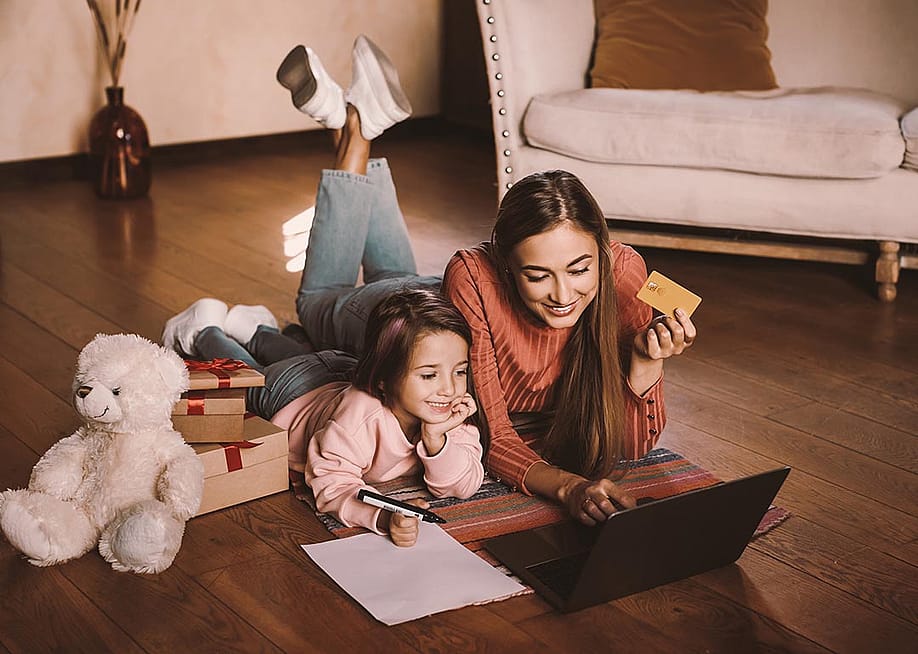 Girl writing wishlist, next to adult woman, both are laying on a rug looking a laptop online shopping while the woman is holding a credit card. There are wrapped gifts next to them along with a teddy bear. — Prostock-studio // Shutterstock