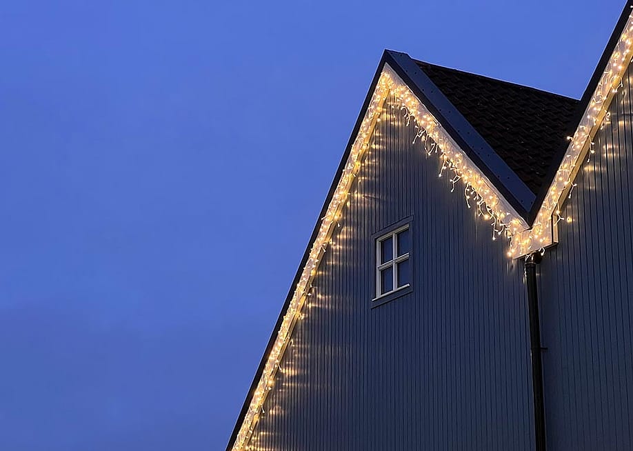Icicle Christmas Lights hanging along the roof line of a wooden-fronted house. There is a small white window on the house, and a dusky blue sky behind the house. — Fraser at Sea // Shutterstock