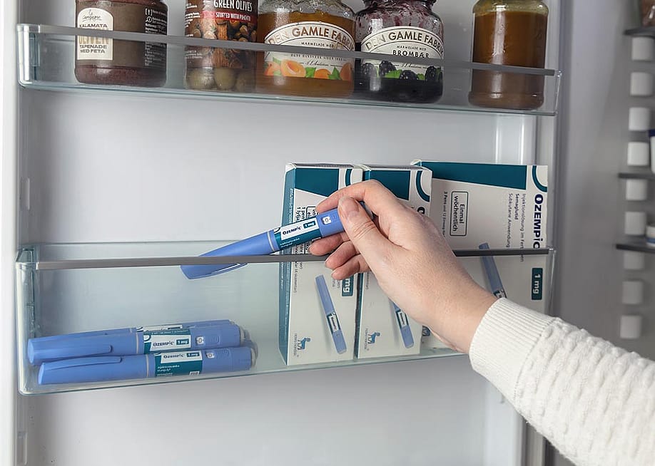 Close up of arm of person wearing white sweater reaching into refrigerator door to pick up an Ozempic injection pen, boxes of Ozempic are stored to the right. — Caroline Ruda // Shutterstock