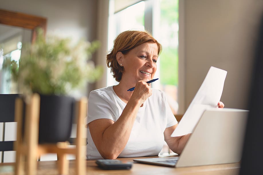 Middle-aged woman sitting at a table looking over paperwork with laptop open and pen in hand. — Lopolo / Shutterstock
