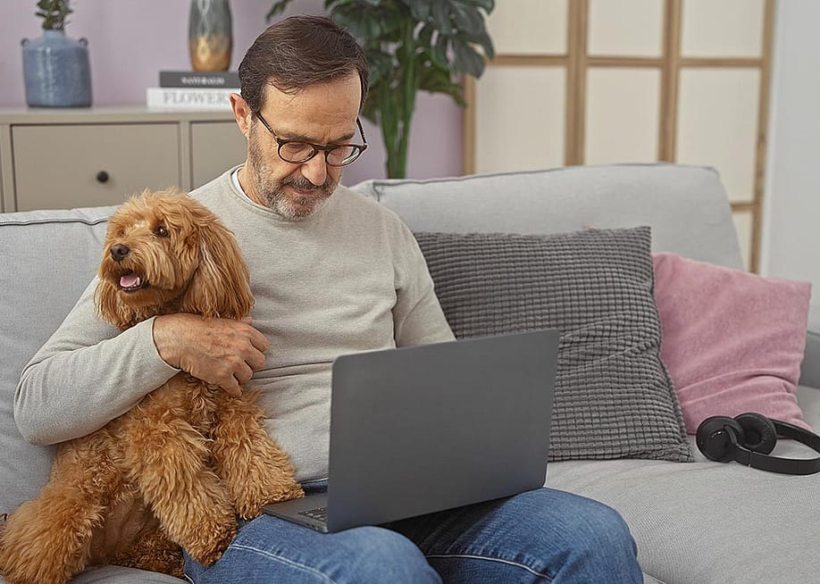 A hispanic middle-aged man cuddles a poodle while using a laptop in a cozy living room setting. — Krakenimages.com // Shutterstock