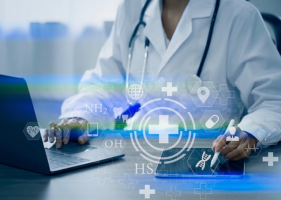 Doctor visible from the neck down sitting at desk with laptop and iPad working with digital medical interface icons superimposed symbolizing AI. — SOMKID THONGDEE // Shutterstock