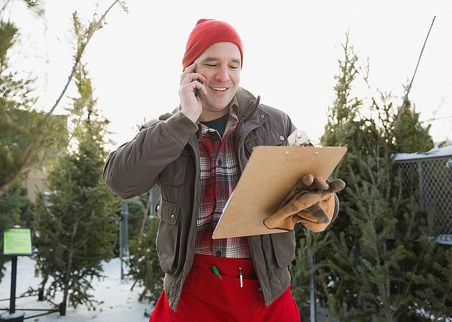 A man talking with someone on the phone while working outdoors for a Christmas tree shop. — Hero Images Inc // Shutterstock