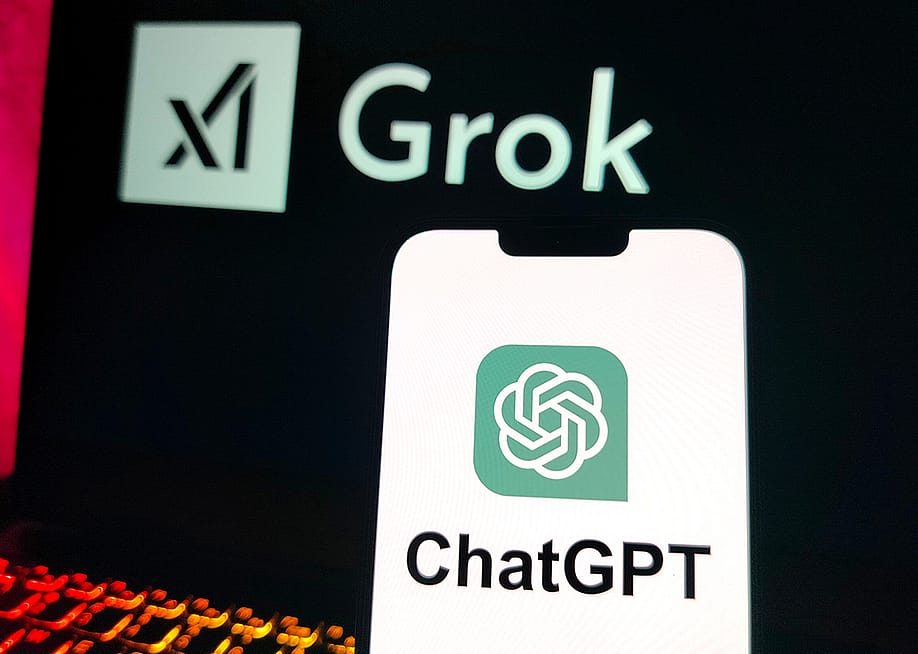 A smartphone displaying the ChatGPT logo and a laptop screen displaying X's Grok logo in the background. — Stock all // Shutterstock