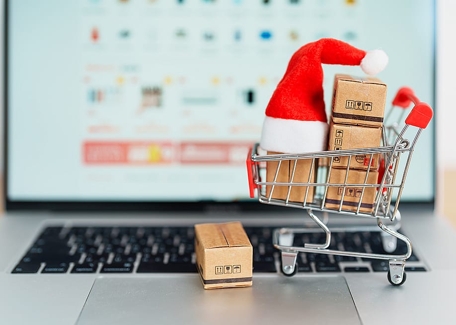 A small shopping cart figure with boxes and a Christmas hat, in front of a laptop displaying a shopping page. — Jo Panuwat D // Shutterstock