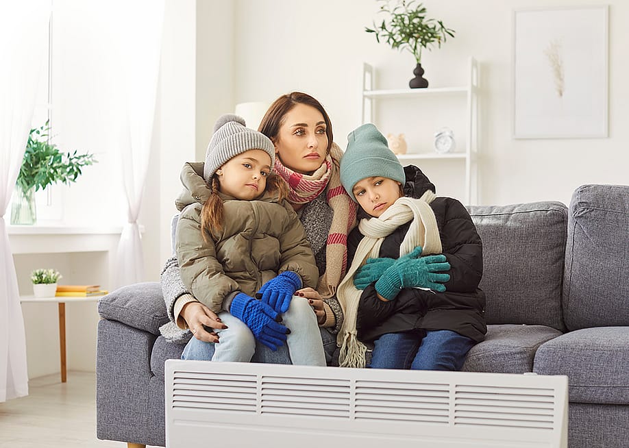 A mother with her two children in winter clothing sitting on a sofa in a cold living room. — Studio Romantic // Shutterstock