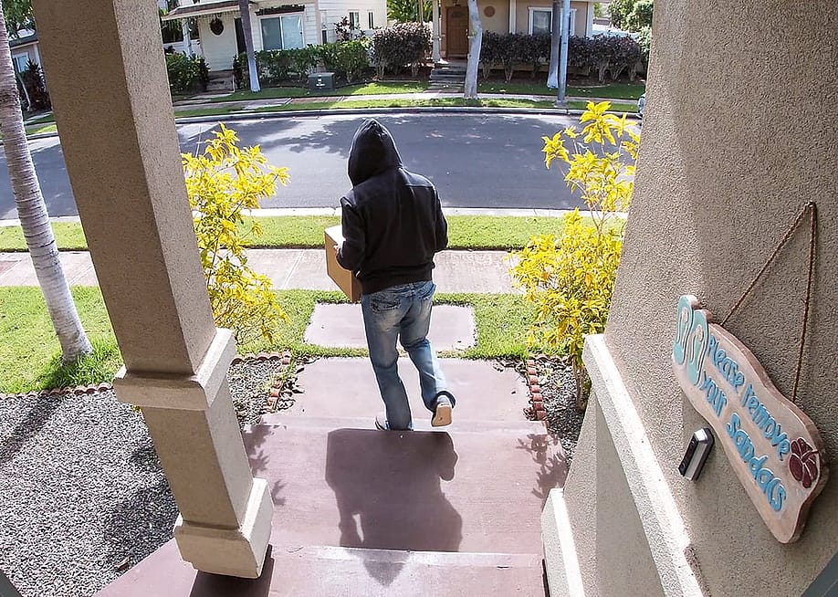 A surveillance camera-view of a package thief walking away from a porch after taking a package. — RightFramePhotoVideo // Shutterstock