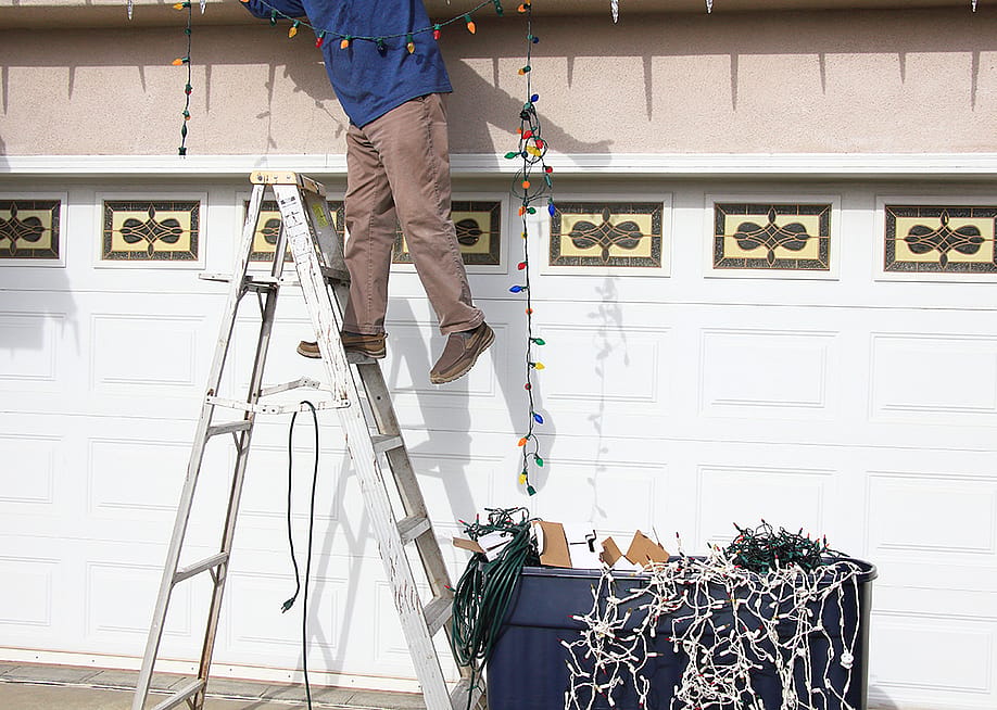A man on a ladder carefully removes Christmas lights in their home. — Wayne Via // Shutterstock