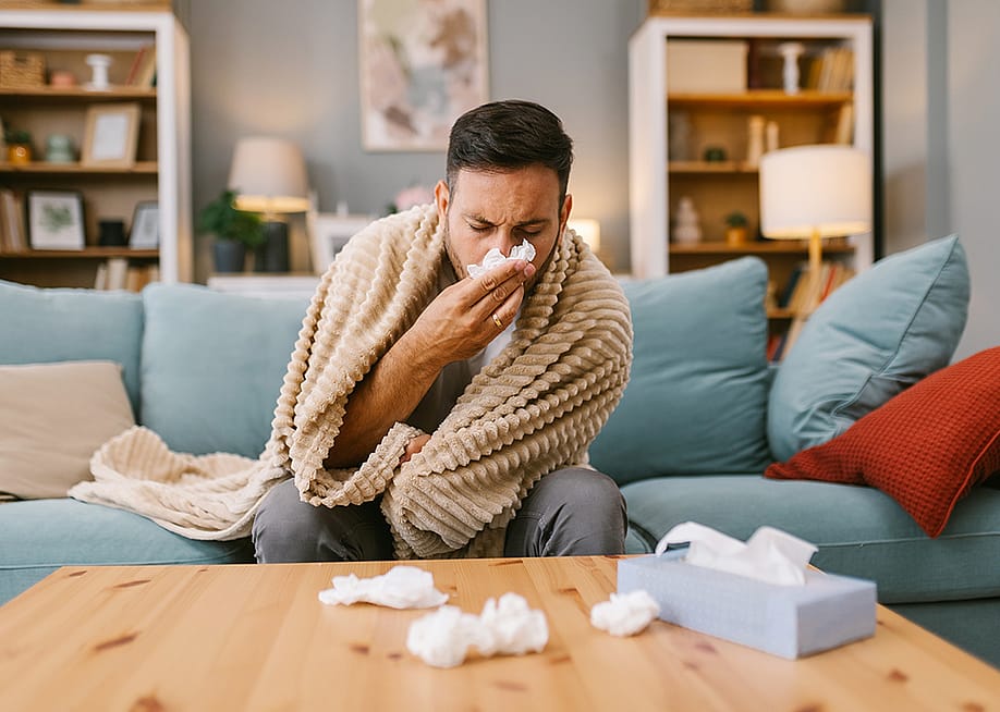 A sick man at home wrapped in blanket sitting on a sofa and blowing his nose. — Cira123 // Shutterstoc