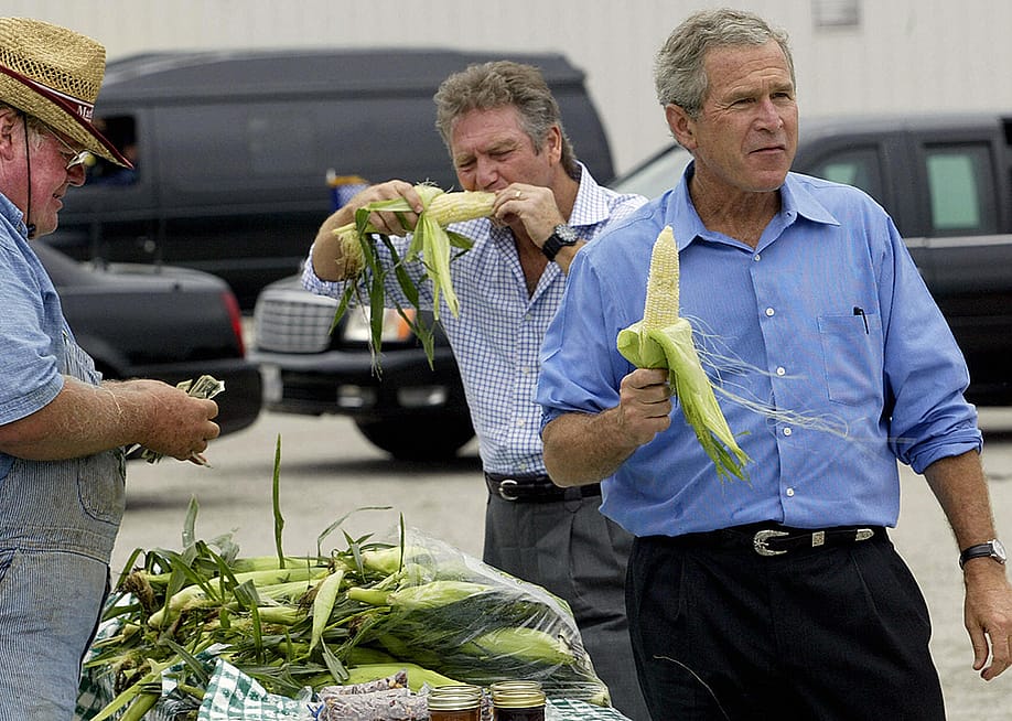 Former US President George W. Bush (R) holding an ear of corn, joined by his friend, Larry Gaitlin (C) while farmer Ken Thompson (L) counts the money handed to him by Bush for a bag of corn, during an impromptu stop of his motorcade at a local farmers market after a campaign rally August 4, 2004 in Davenport, Iowa. — Tim Sloan // AFP via Getty Images