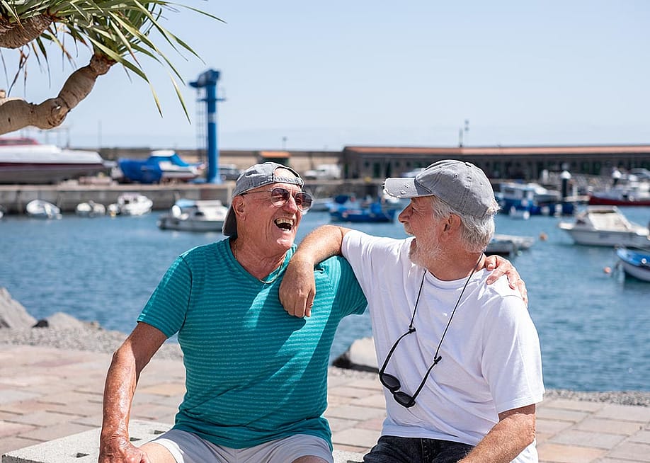 Two senior friends wearing caps and t-shirts in retirement life sitting on a bench at the harbor looking each other laughing happily. — Lucigerma // Shutterstock.