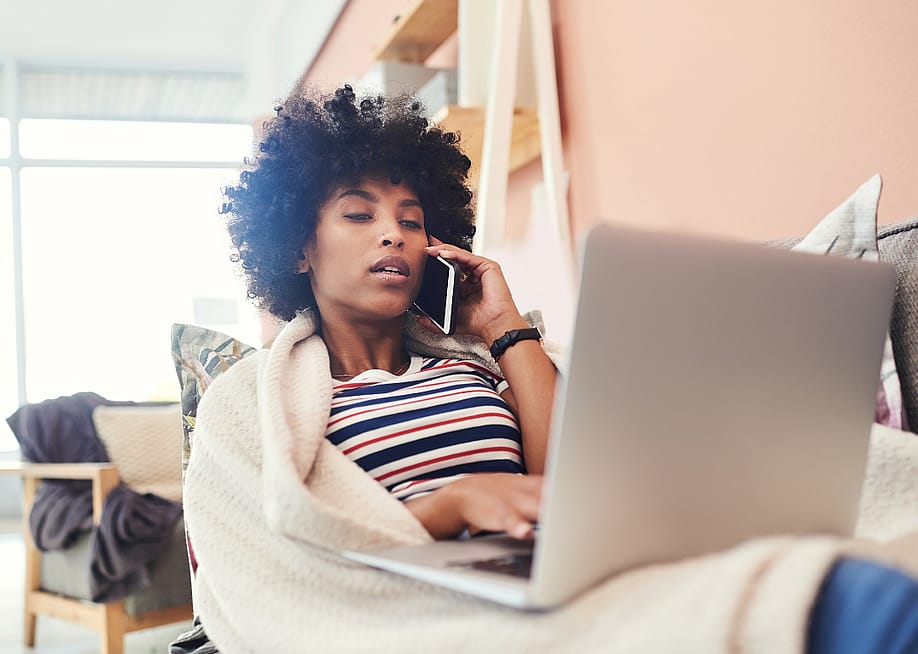A woman at home on the couch, wrapped in a blanket talking on the phone with a laptop on her lap. — PeopleImages // Shutterstock