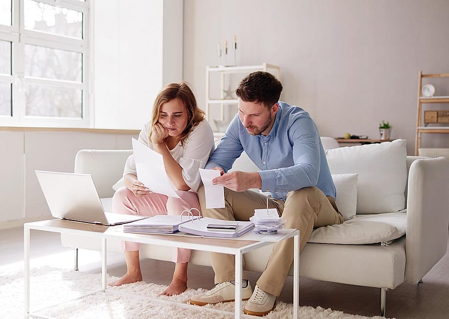 Two people sitting on a white sofa looking at bills and paperwork in a minimalistic living room. — Andrey_Popov // Shutterstock