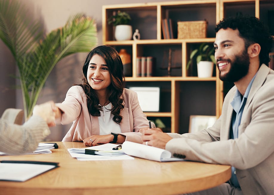 Three business people sit at a table. One smiling person shakes hands with another person, who is mostly out of view. — PeopleImages // Shutterstock