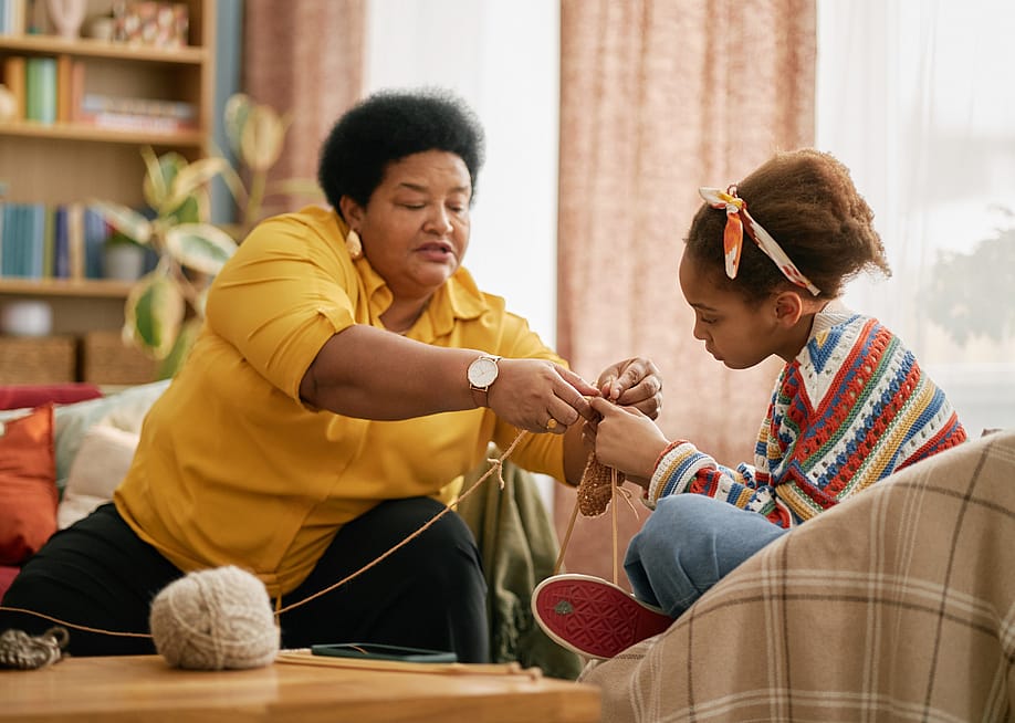 An older woman and a young girl sit in a living room as they wind yarn. — SeventyFour // Shutterstock