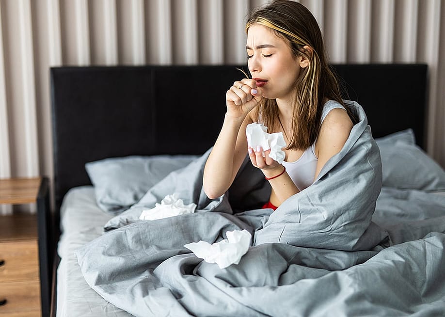 A sick woman wrapped in blanket is sitting on her bed and coughing. — F8 studio // Shutterstock