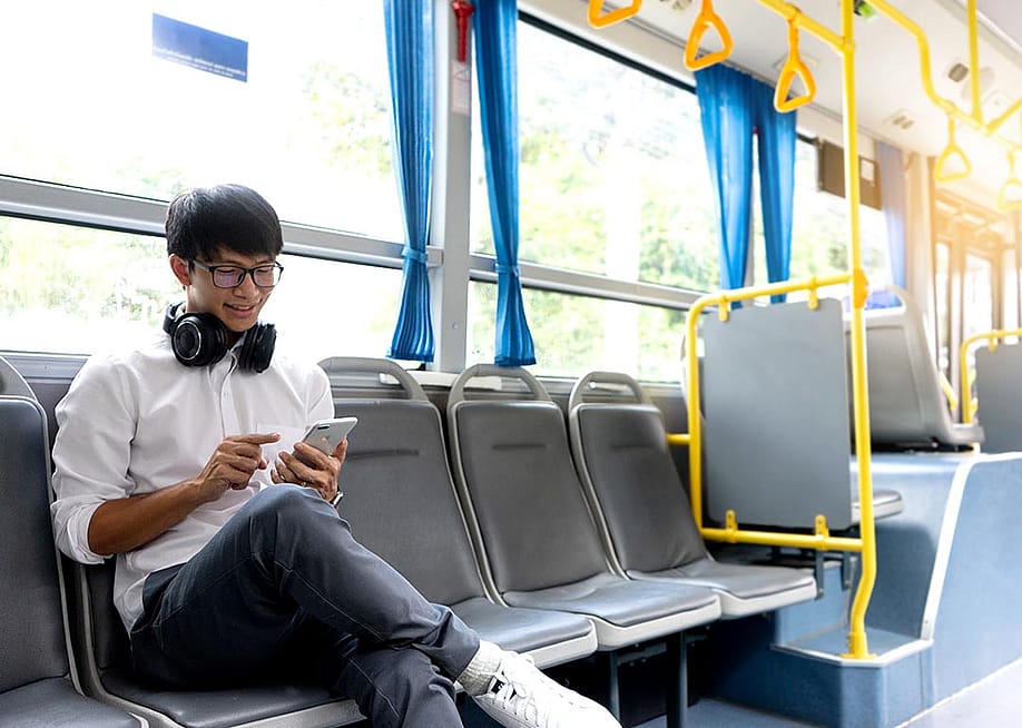 A college-age person wearing a crisp white shirt and headphones around his neck and bright white shoes sits on the side-facing seats in public transportation looking attentively at phone. — sabthai // Shutterstock