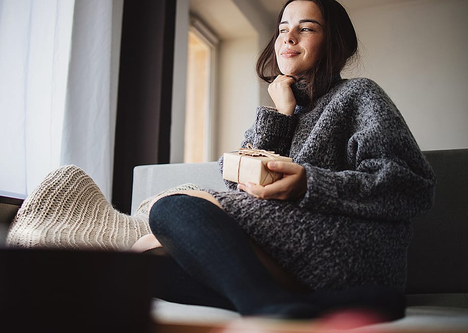 A young woman cozily sitting on a sofa at home holding a small gift box. — Flotsam // Shutterstock
