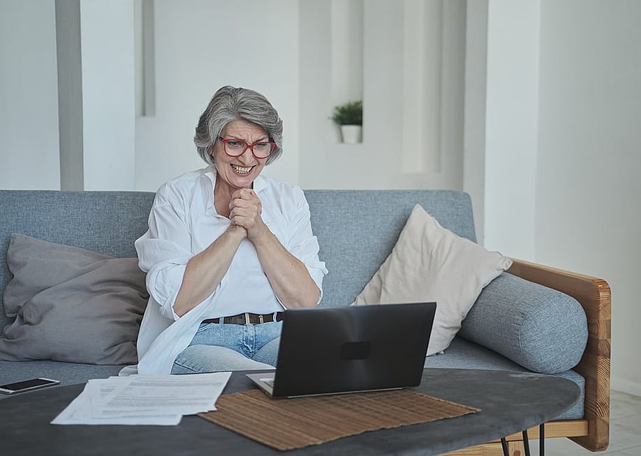 A senior woman sitting on a sofa and excitingly awaits good news on her laptop. — Justlight // Shutterstock