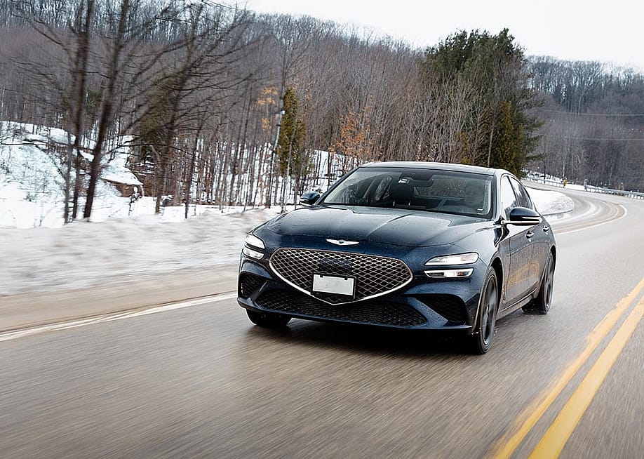 Front view of black Genesis G70 driving on a road in winter with snow and trees in background. — Genesis