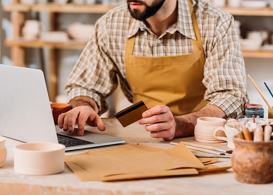 Cropped view of a small pottery business owner holding a credit card and browsing a laptop. — LightField Studios // Shutterstock