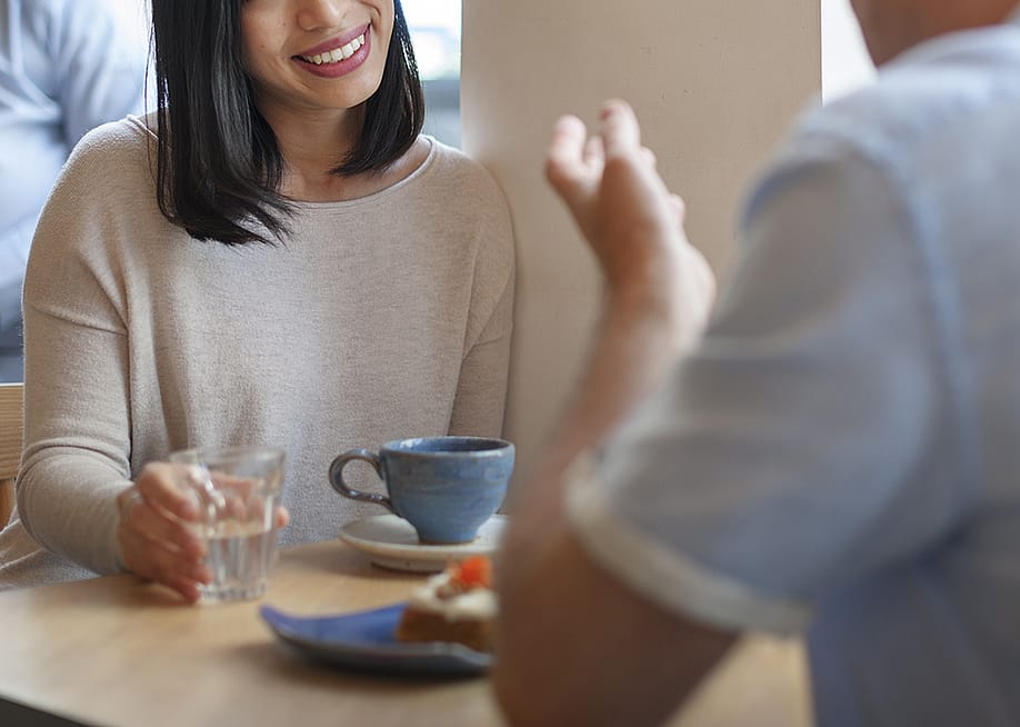 Two people sitting across from each other at a table in a cafe with water, a piece of cake, a mug in front of them. Just one person's smiling face is partially in view. — Rawpixel.com // Shutterstock
