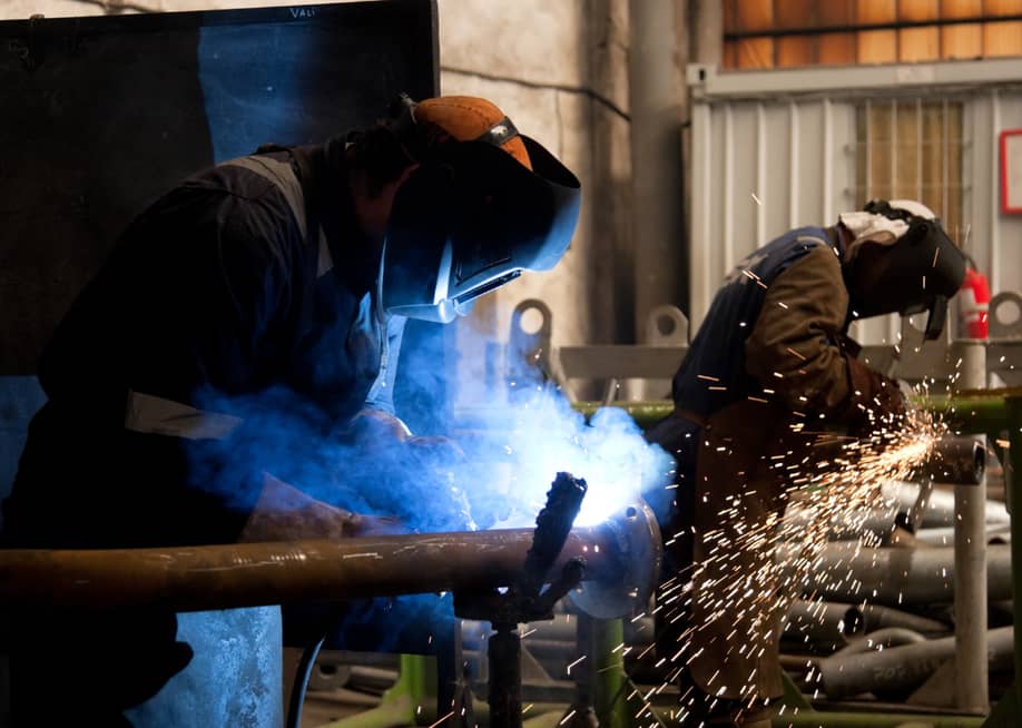 Two welders with protective masks welding metal, with sparks flying around. — Bogdan VASILESCU // Shutterstock