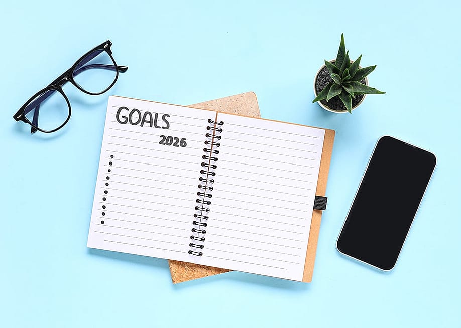 A pair of eyeglasses, a smartphone, and a notebook to list 2026 goals with a blue background. — Pixel-Shot // Shutterstock