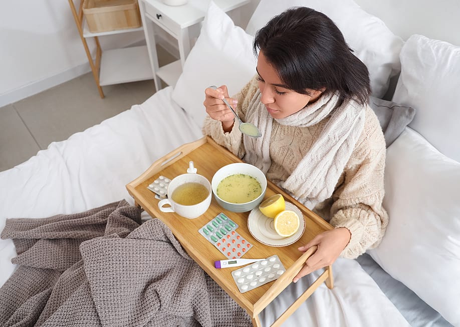 A woman sits in bed holding a spoon full of soup with a tray over her lap containing pills, nasal spray, a thermometer, lemons, a bowl of soup, and a mug of tea. — Pixel-Shot // Shutterstock