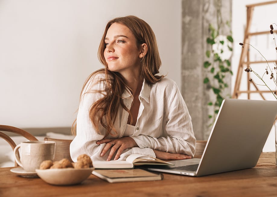 A content-looking woman in her 20s or 30s sits in a sunny room at a table with a laptop, a mug, a notebook, and a bowl of baked goods. — Dean Drobot // Shutterstock