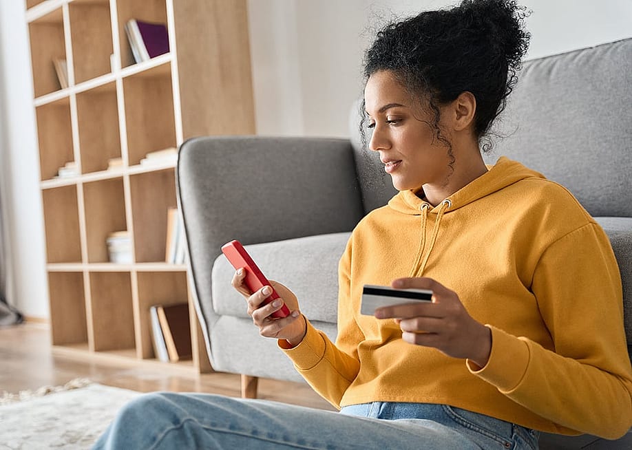 A young woman sitting on the floor of a living room, holding a credit card and browsing her smartphone. — Ground Picture // Shutterstock