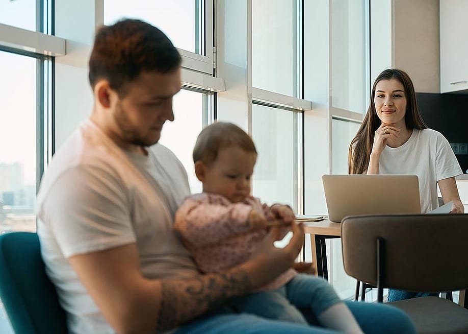 Woman smiling and sitting at table with laptop looking at man holding baby in the foreground. — YAKOBCHUK VIACHESLAV // Shutterstock.
