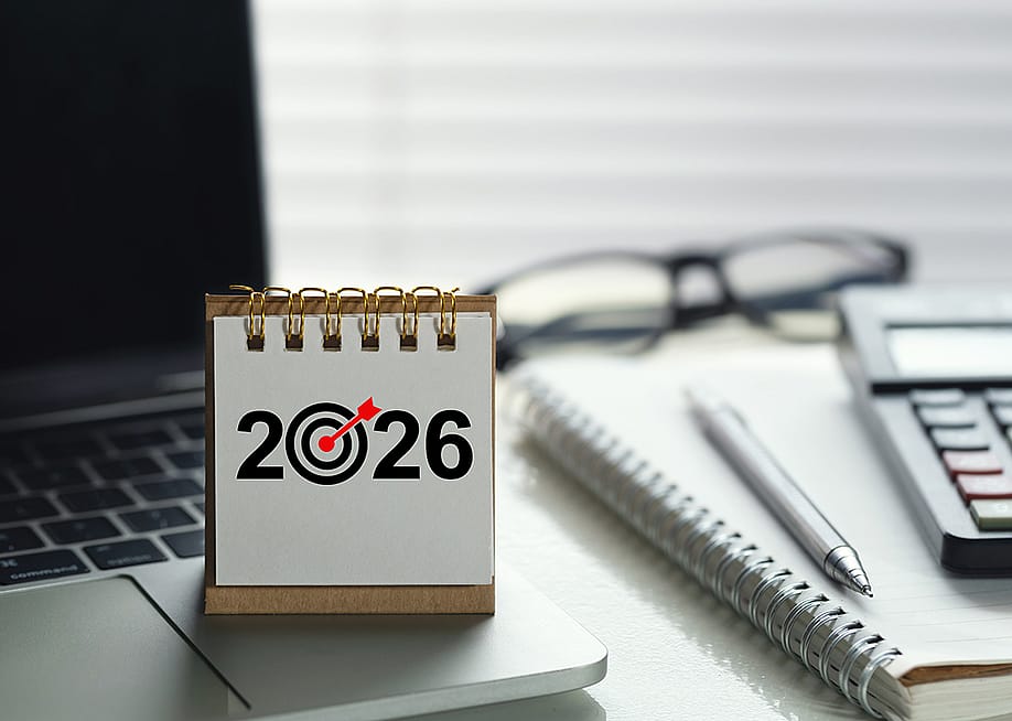 A desk calendar for 2026 with a target icon on top of a laptop among other office desk items. — chayanuphol // Shutterstock