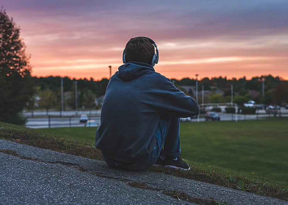 Rear view of a male teenager sitting alone at the top of a hill and watching sunset while listening to headphones. — mooremedia // Shutterstock
