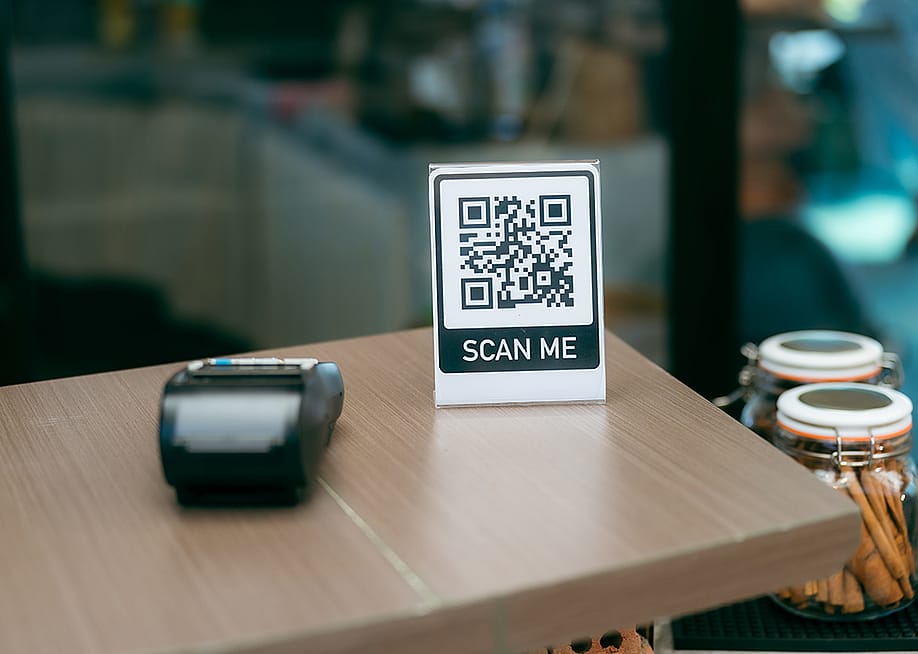 A 'scan me' QR code sign and payment terminal on a cafe reception table. — Chokniti-Studio // Shutterstock