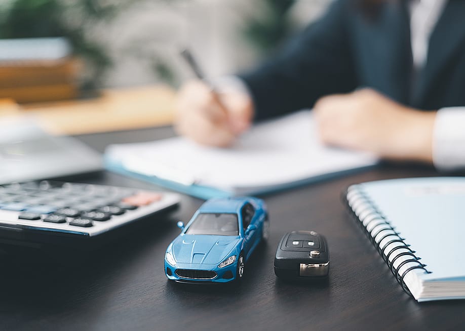 A small, blue car figure beside a car key in front of a car agent reviewing contracts. — jd8 // Shutterstock