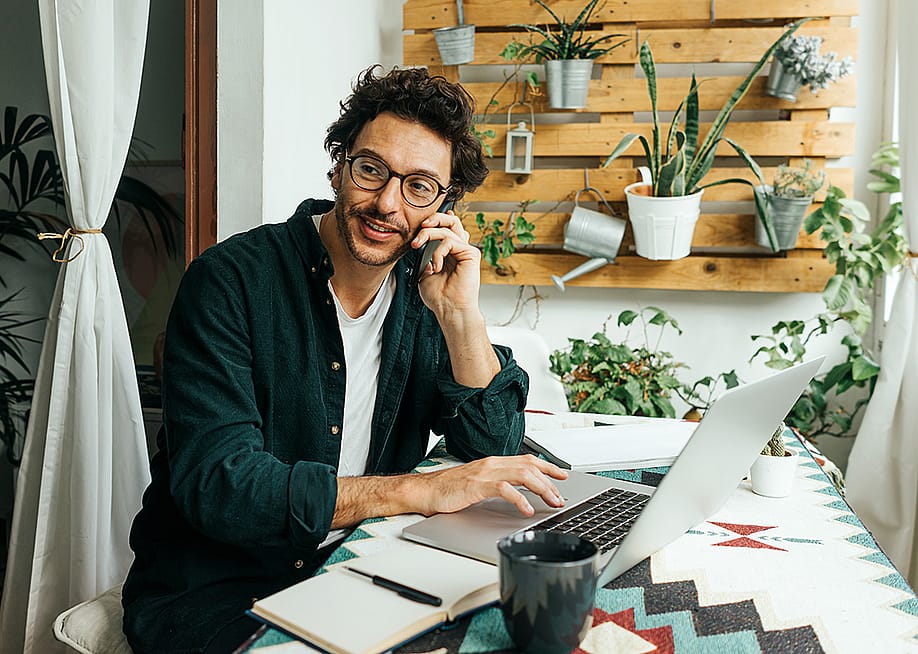 A young man on phone call and using laptop to work remotely from home. — Rymden // Shutterstock
