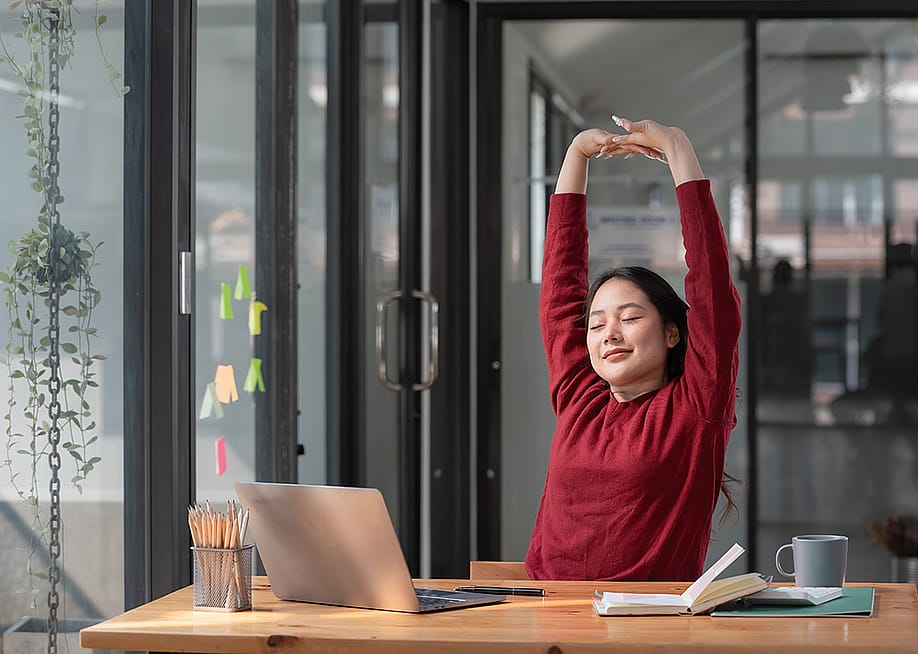 A young female employee stretching up her hands while working in the office. — Songsak C // Shutterstock