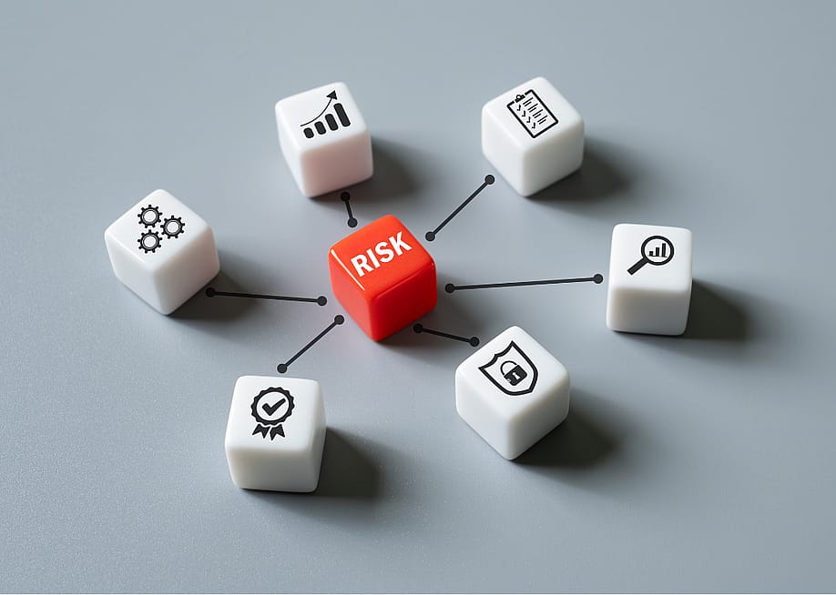 One red die with the word risk on it, positioned in in the middle of six white dice with symbols, including three cogs, an upward bar graph, a checklist, a magnifying glass over a rising bar graph, a security shield with a lock in the middle, and a ribbon with a check mark. — BritCats Studio // Shutterstock