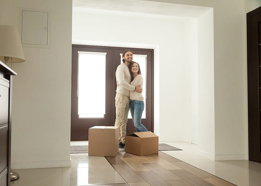 A hugging couple stand in the house in front of the door with two cardboard boxes in front of them. — fizkes // Shutterstock