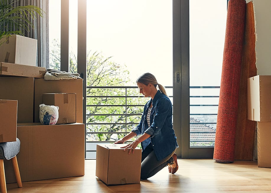 A women kneels in front of a cardboard box in a bright room with other boxes and household items like lamps and furniture. — PeopleImages // Shutterstock