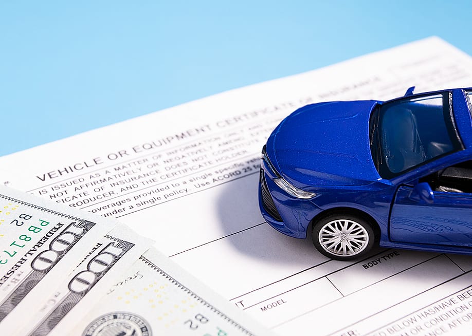 A small figure of a blue car on top of banknotes and insurance documents. — Yta23 // Shutterstock