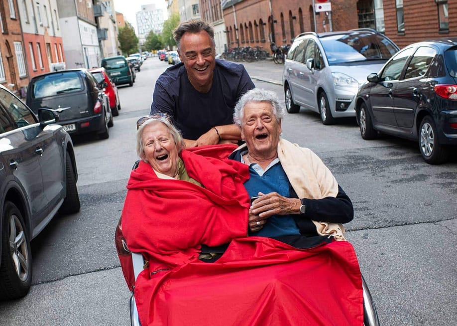 A volunteer cycles with a trishaw carrying two seated passengers who are laughing and smiling. — Courtesy of Cycling Without Age